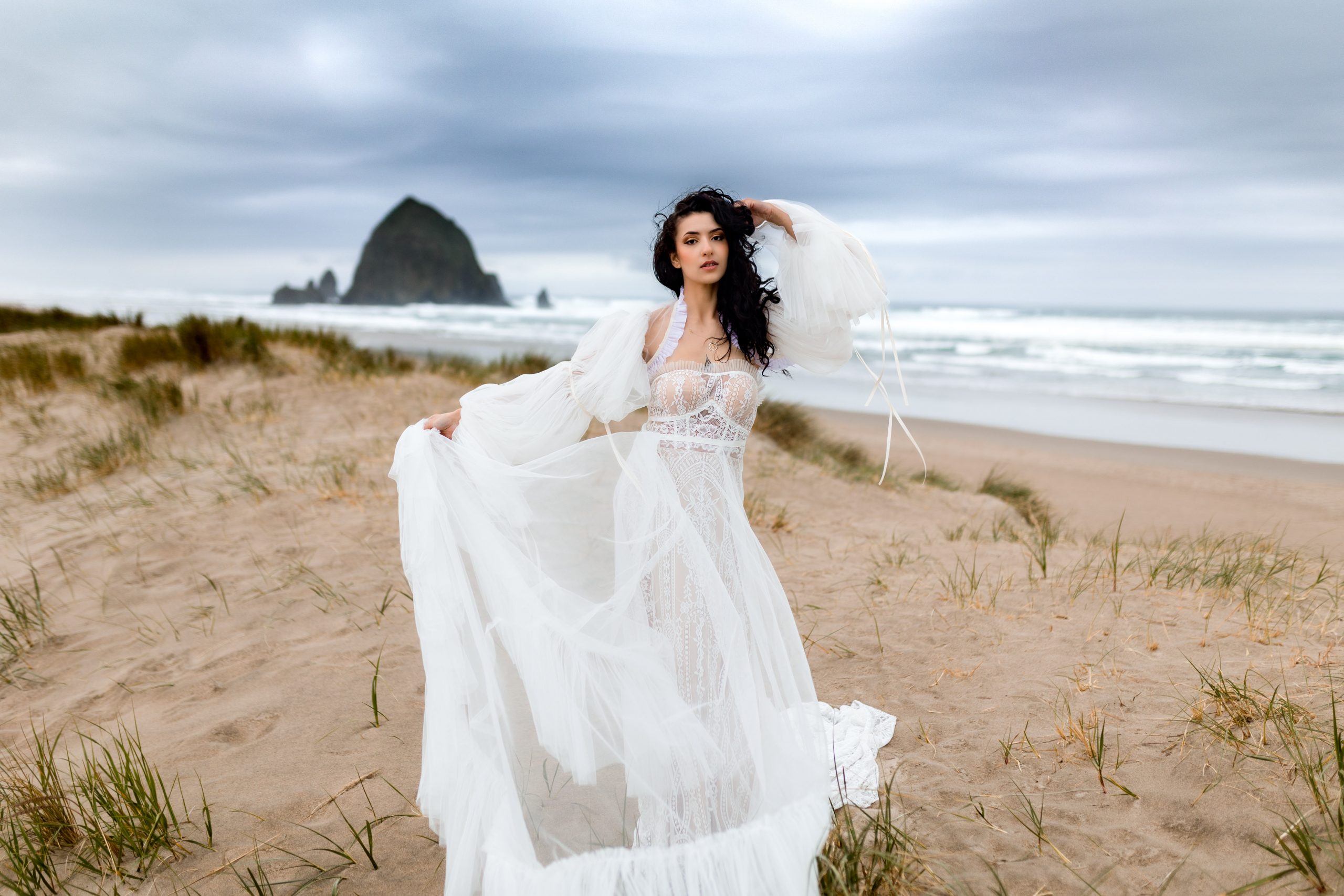 A bride swaying in the breeze holding her wedding dress train on Cannon Beach, Oregon with the rock stacks behind her