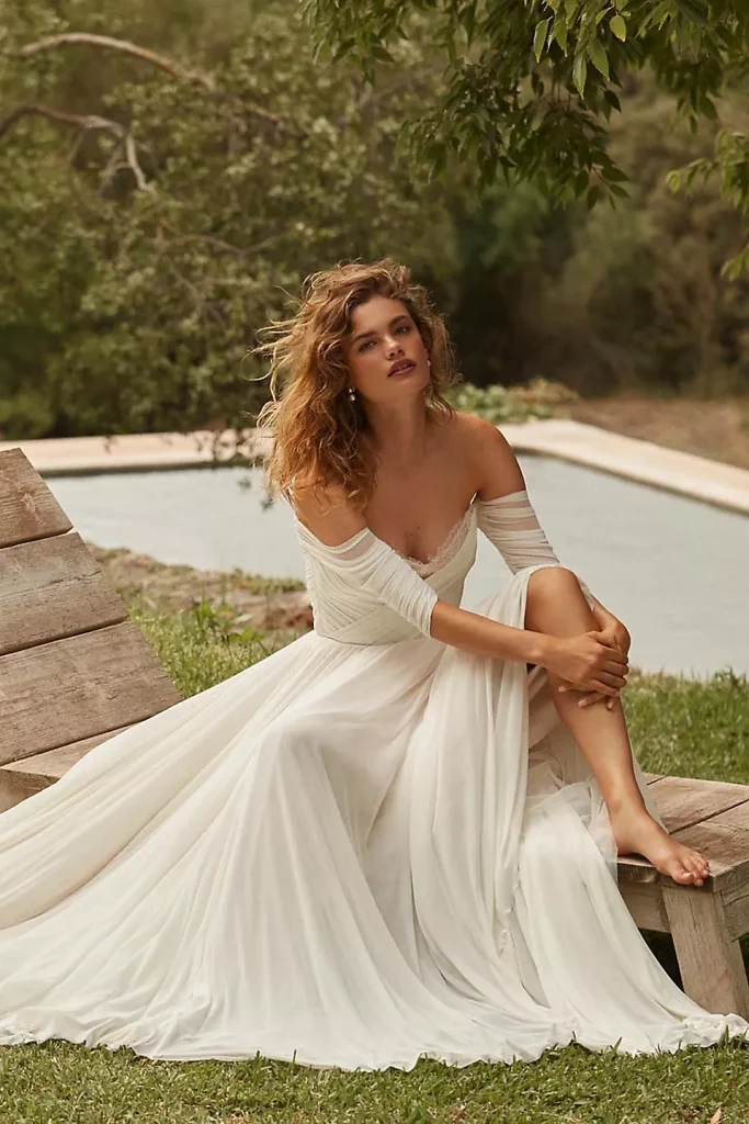 A bride sitting on a wooden lounge chair near a small pool in a gorgeous mesh off-the-shoulder wedding gown, hugging her knee and looking at the camera