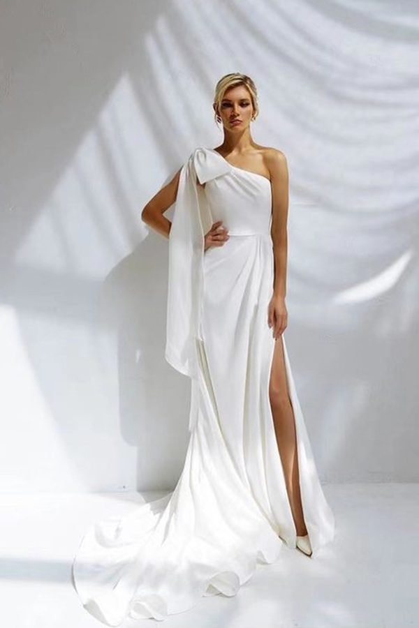 a bride standing in front of a white sheet background posing in a crepe wedding dress that features a one shoulder ribbon